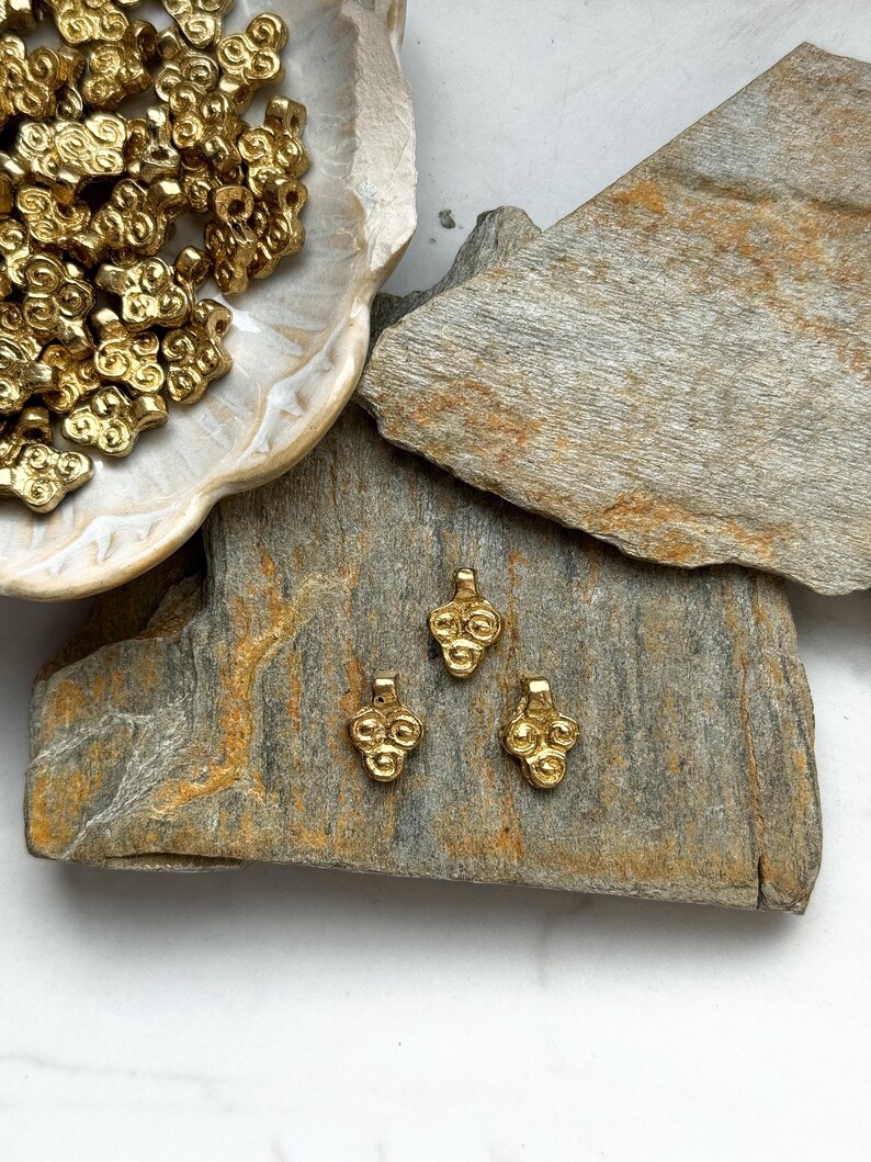 May include: A close-up of three gold-colored metal charms shaped like a cluster of grapes. The charms are sitting on a piece of rough, gray stone.