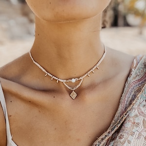 May include: A delicate layered necklace with a small gold pendant featuring a geometric design. The necklace is made of white beads and has gold accents.