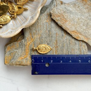 May include: Gold leaf charm pendant with a detailed design. The charm is approximately 1 inch long and is shown next to a ruler for size reference.