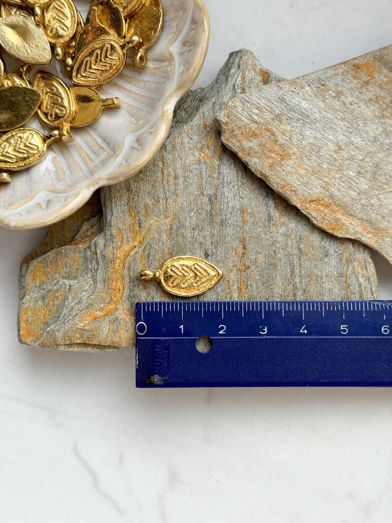 May include: Gold leaf charm pendant with a detailed design. The charm is approximately 1 inch long and is shown next to a ruler for size reference.