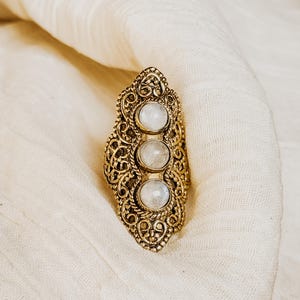Puede incluir: Anillo dorado ornamentado con tres piedras ovaladas de color blanco lechoso. El anillo tiene un intrincado diseño de filigrana y está sobre un fondo de tela color crema. Una pieza de joyería llamativa.
