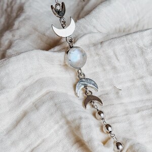 May include: A silver-toned chain necklace featuring a series of metal moon phase charms and small spherical beads. The centerpiece is a round, iridescent white gemstone set in a silver-colored bezel, flanked by crescent moon shapes.