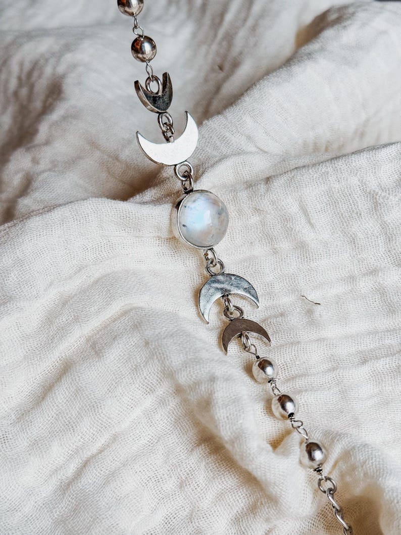 May include: A silver-toned chain necklace featuring a series of metal moon phase charms and small spherical beads. The centerpiece is a round, iridescent white gemstone set in a silver-colored bezel, flanked by crescent moon shapes.