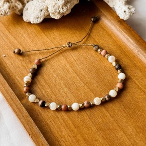 May include: A beaded bracelet featuring a mix of round white, brown, and pink beads interspersed with small, faceted metallic gold-toned spacers. The bracelet is constructed with a thin brown cord and includes an adjustable sliding knot closure.