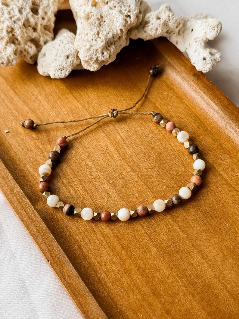 May include: A beaded bracelet featuring a mix of round white, brown, and pink beads interspersed with small, faceted metallic gold-toned spacers. The bracelet is constructed with a thin brown cord and includes an adjustable sliding knot closure.