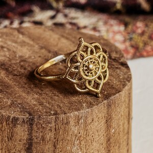 K&ouml;nnte beinhalten: Ein goldfarbener Ring mit einem filigranen, komplizierten Design, das an eine Blume erinnert. Der Ring liegt auf einer Holzoberfl&auml;che.