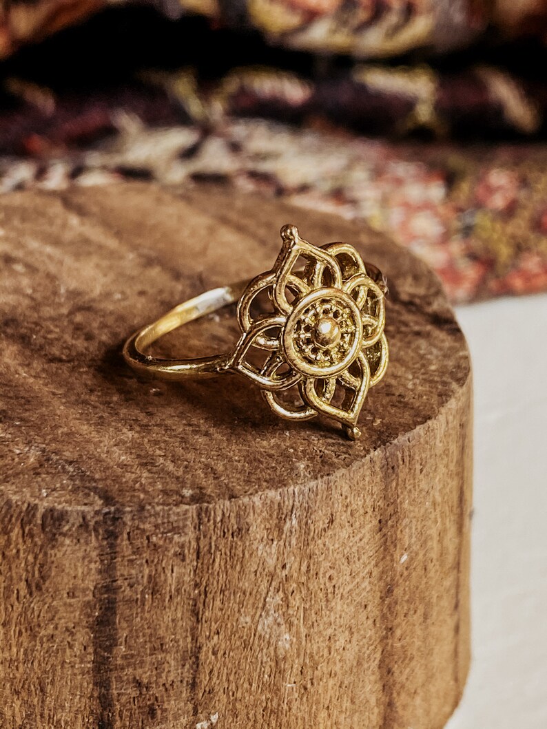 K&ouml;nnte beinhalten: Ein goldfarbener Ring mit einem filigranen, komplizierten Design, das an eine Blume erinnert. Der Ring liegt auf einer Holzoberfl&auml;che.