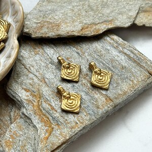 K&ouml;nnte beinhalten: Drei goldfarbene Metall-Anh&auml;nger mit quadratischer Form und einem Spiralmuster in der Mitte. Die Anh&auml;nger liegen auf einer rauen, braunen, strukturierten Oberfl&auml;che.
