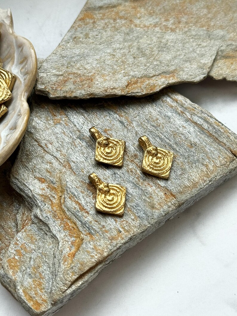 K&ouml;nnte beinhalten: Drei goldfarbene Metall-Anh&auml;nger mit quadratischer Form und einem Spiralmuster in der Mitte. Die Anh&auml;nger liegen auf einer rauen, braunen, strukturierten Oberfl&auml;che.