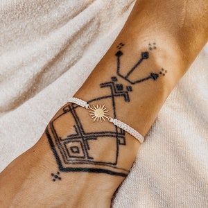 May include: A white string bracelet with a gold sun charm. The bracelet is on a person's wrist with a black tattoo.
