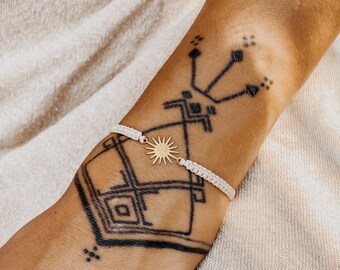 May include: A white string bracelet with a gold sun charm. The bracelet is on a person's wrist with a black tattoo.