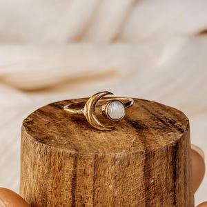 Puede incluir: Un anillo de oro en forma de media luna con una piedra de ópalo blanco. El anillo está en un expositor de madera.