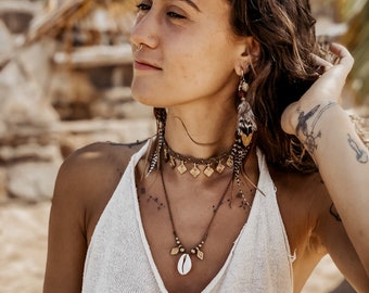 May include: A woman with long brown hair wearing a white tank top and multiple gold necklaces with a shell pendant. She is wearing a nose ring and feather earrings.