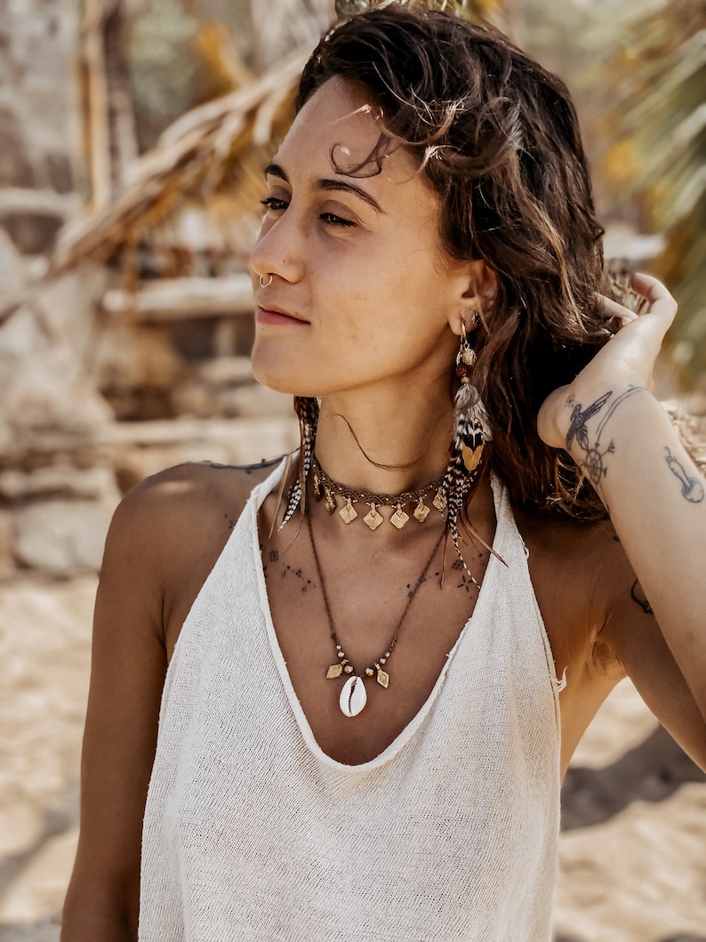 May include: A woman with long brown hair wearing a white tank top and multiple gold necklaces with a shell pendant. She is wearing a nose ring and feather earrings.