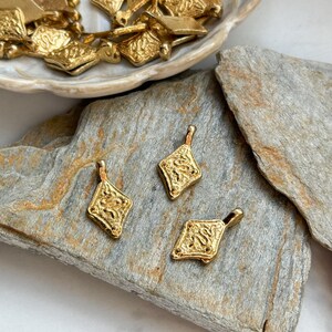 Pode incluir: Pingentes dourados em forma de losango com designs ornamentados e texturizados. Tr&ecirc;s pingentes s&atilde;o dispostos sobre uma superf&iacute;cie de pedra cinza, com pingentes adicionais em um prato branco no fundo. Os pingentes t&ecirc;m uma argola na parte superior para enfiar.