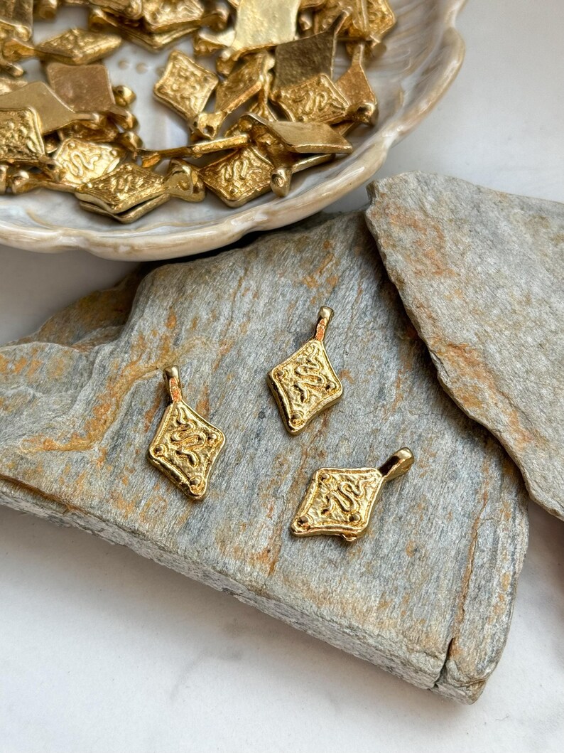Pode incluir: Pingentes dourados em forma de losango com designs ornamentados e texturizados. Tr&ecirc;s pingentes s&atilde;o dispostos sobre uma superf&iacute;cie de pedra cinza, com pingentes adicionais em um prato branco no fundo. Os pingentes t&ecirc;m uma argola na parte superior para enfiar.