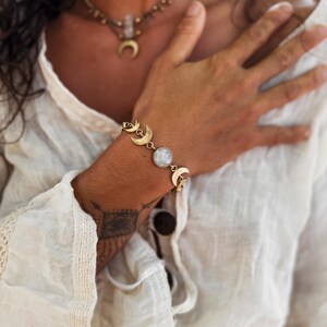 May include: A gold-toned bracelet featuring a series of crescent moon charms and a central round, iridescent white stone. The bracelet is worn on a wrist, accompanied by a matching gold-toned necklace with a crescent moon pendant.