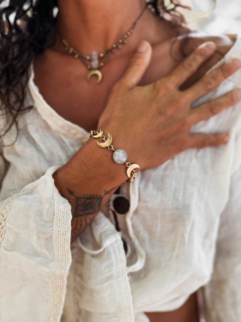 May include: A gold-toned bracelet featuring a series of crescent moon charms and a central round, iridescent white stone. The bracelet is worn on a wrist, accompanied by a matching gold-toned necklace with a crescent moon pendant.
