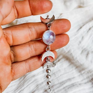 May include: A silver-toned bracelet featuring a central round iridescent moonstone cabochon flanked by two silver crescent moon charms. The chain continues with a series of small silver spheres and standard links, ending in a metal clasp.