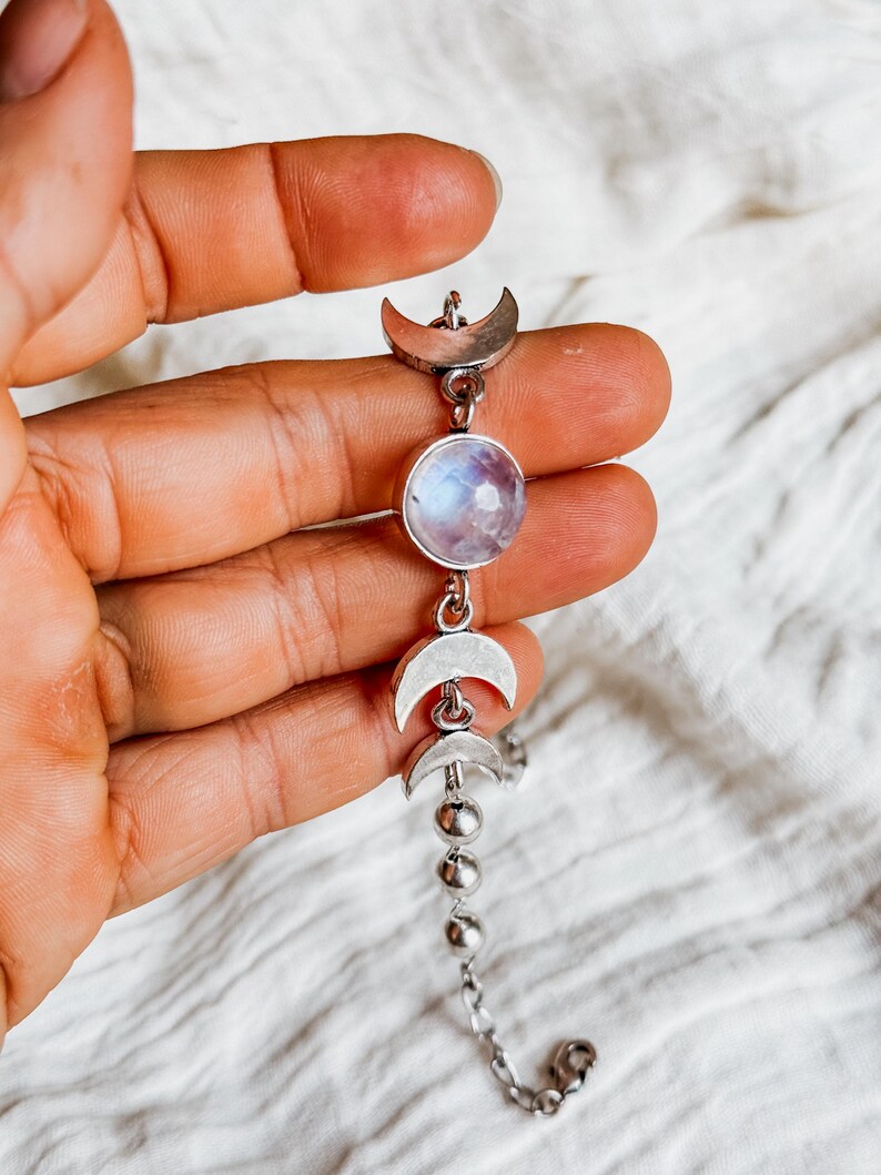May include: A silver-toned bracelet featuring a central round iridescent moonstone cabochon flanked by two silver crescent moon charms. The chain continues with a series of small silver spheres and standard links, ending in a metal clasp.