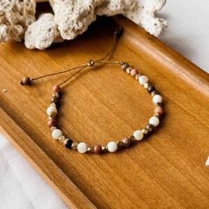 May include: A beaded bracelet featuring a repeating pattern of white, brown, and pink spherical beads interspersed with small, faceted metallic gold-toned spacers. The bracelet is strung on a thin brown cord with an adjustable sliding knot closure.