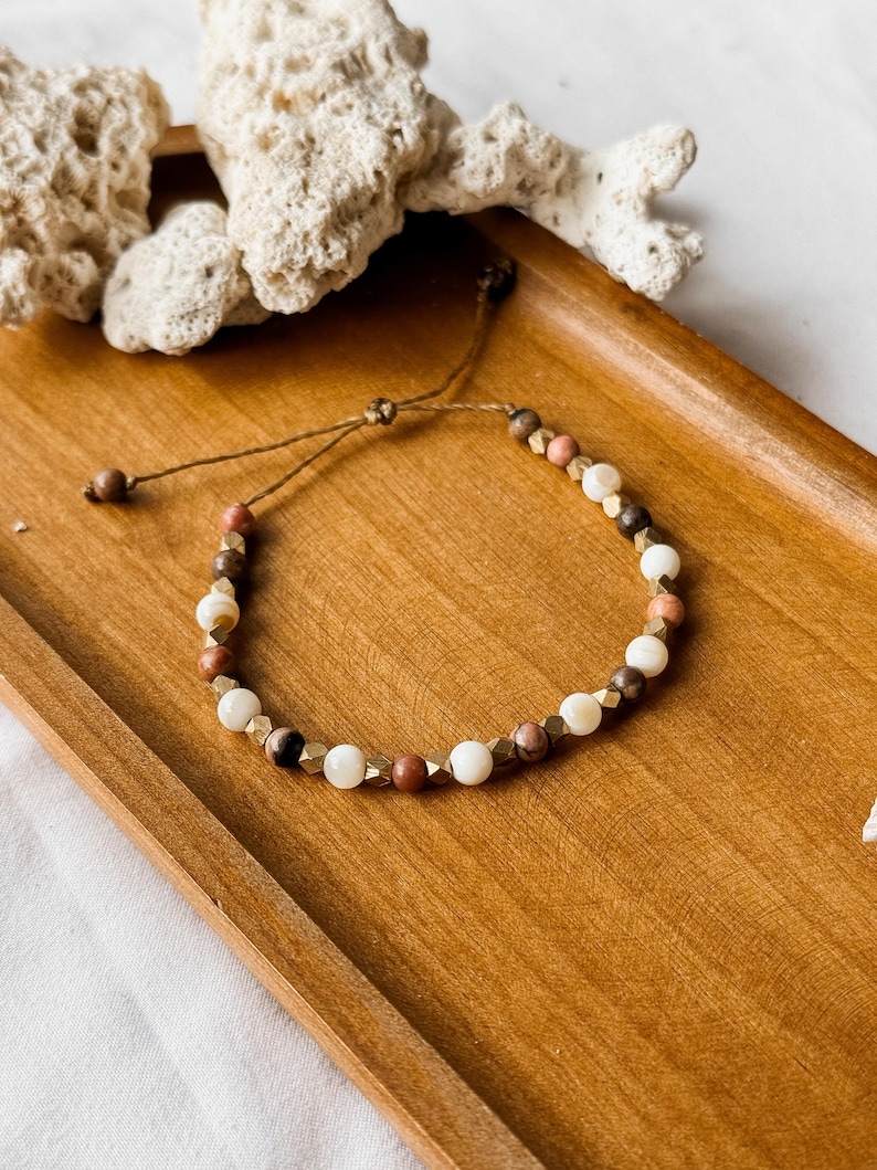 May include: A beaded bracelet featuring a repeating pattern of white, brown, and pink spherical beads interspersed with small, faceted metallic gold-toned spacers. The bracelet is strung on a thin brown cord with an adjustable sliding knot closure.