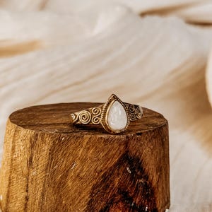 Puede incluir: Un anillo de oro con una piedra preciosa blanca en forma de pera engastada en el centro. La banda del anillo está decorada con un patrón de espiral.