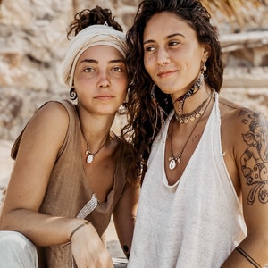 May include: Two women wearing bohemian style clothing and jewelry. One woman is wearing a brown tank top with a shell necklace and a white head wrap. The other woman is wearing a white tank top with a shell necklace and multiple bracelets. Both women have long hair and are smiling.