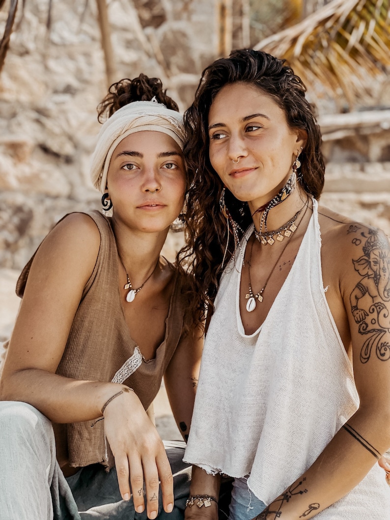 May include: Two women wearing bohemian style clothing and jewelry. One woman is wearing a brown tank top with a shell necklace and a white head wrap. The other woman is wearing a white tank top with a shell necklace and multiple bracelets. Both women have long hair and are smiling.