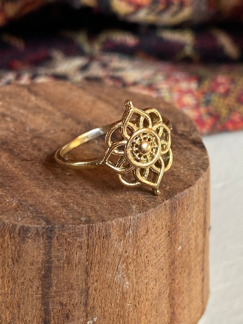 K&ouml;nnte beinhalten: Ein goldfarbener Ring mit einem filigranen, kunstvollen Blumendesign. Der Ring hat eine kreisf&ouml;rmige Mitte mit einem kleinen, erhabenen Punkt.