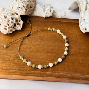 May include: A bracelet featuring a repeating pattern of round white beads, round olive green beads, and small metallic gold-toned faceted beads strung on a thin brown cord. The bracelet includes an adjustable sliding knot closure with two small beads.