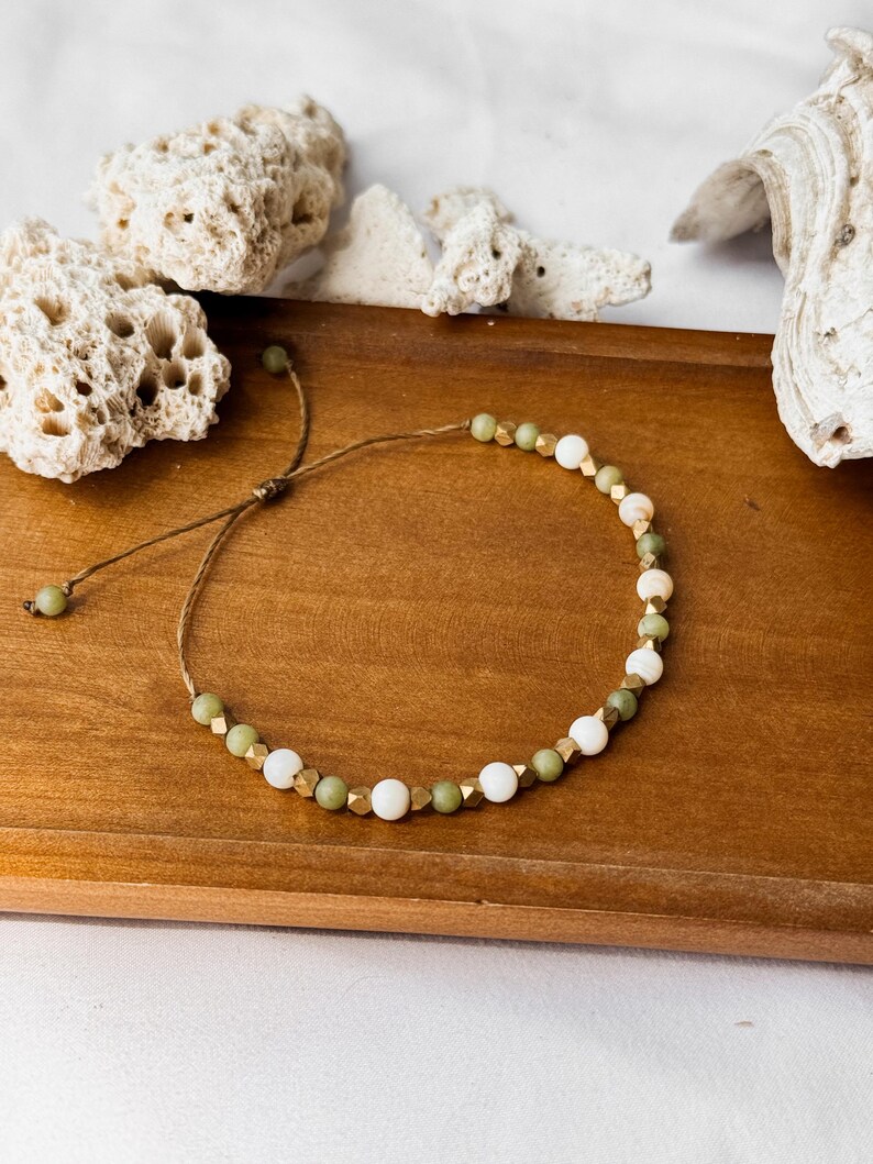 May include: A bracelet featuring a repeating pattern of round white beads, round olive green beads, and small metallic gold-toned faceted beads strung on a thin brown cord. The bracelet includes an adjustable sliding knot closure with two small beads.