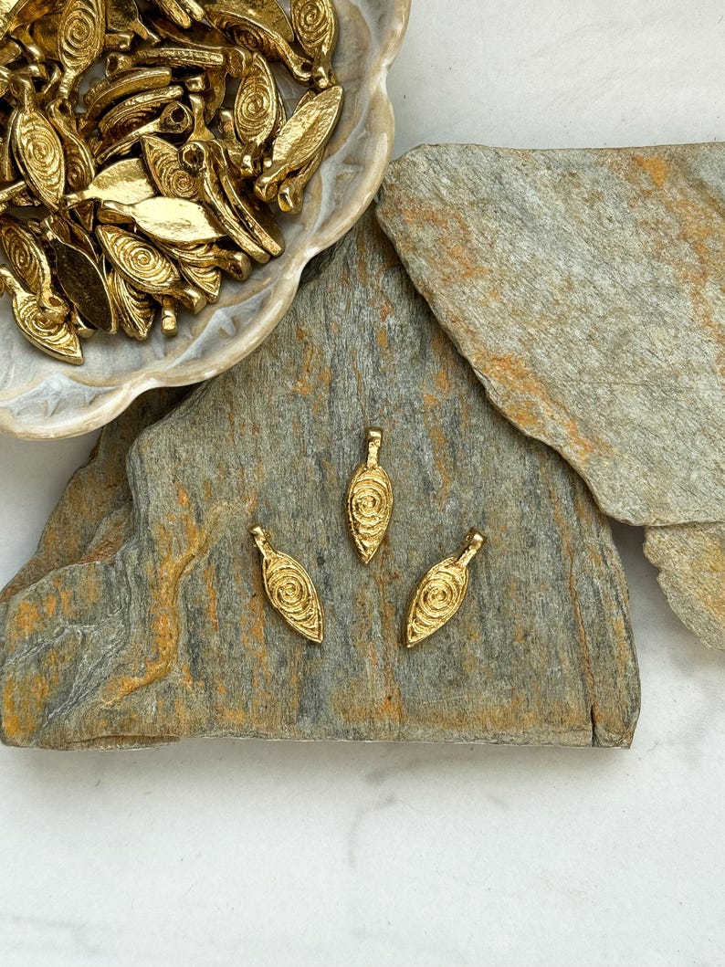 May include: Three gold-colored metal pendants with a swirl design. The pendants are on a gray stone surface.