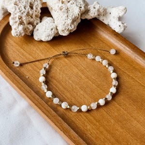 May include: A delicate adjustable bracelet featuring a repeating pattern of round white beads, translucent clear beads, and small metallic gold-toned geometric spacers. The bracelet is secured with a brown cord and a sliding knot closure mechanism.