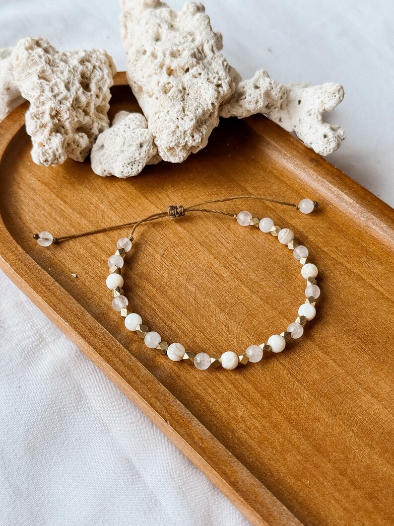 May include: A delicate adjustable bracelet featuring a repeating pattern of round white beads, translucent clear beads, and small metallic gold-toned geometric spacers. The bracelet is secured with a brown cord and a sliding knot closure mechanism.