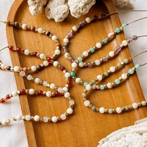 May include: A collection of six beaded bracelets arranged on a wooden tray. Each bracelet features a combination of white beads, metallic gold-toned faceted spacers, and various colored gemstone beads including red, green, and purple hues.