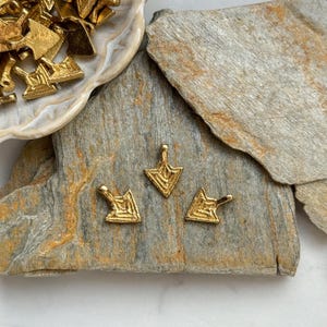 May include: Three gold-colored metal arrow charms with a geometric design. The charms are on a gray and brown stone surface.