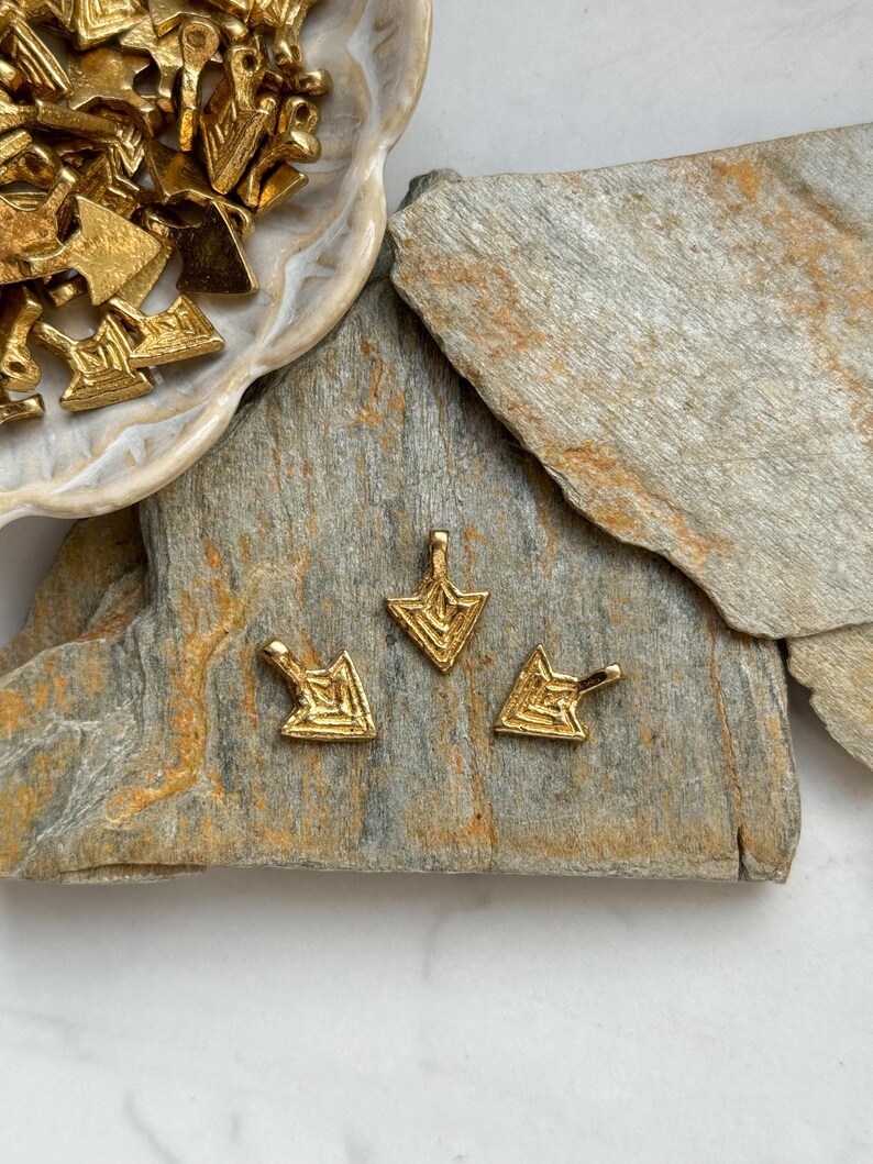 May include: Three gold-colored metal arrow charms with a geometric design. The charms are on a gray and brown stone surface.