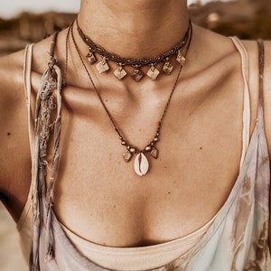May include: A layered necklace with a shell pendant and small gold charms. The necklace is made of brown cord and has a braided choker style.