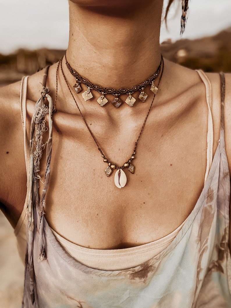 May include: A layered necklace with a shell pendant and small gold charms. The necklace is made of brown cord and has a braided choker style.
