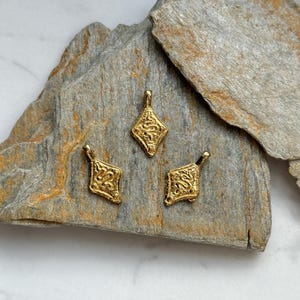 Pode incluir: Tr&ecirc;s pingentes dourados e ornamentados, com formato de losango, s&atilde;o exibidos em uma superf&iacute;cie de pedra cinza. Cada pingente apresenta um design intrincado e texturizado, com uma pequena argola no topo para pendurar. Ideal para joias.