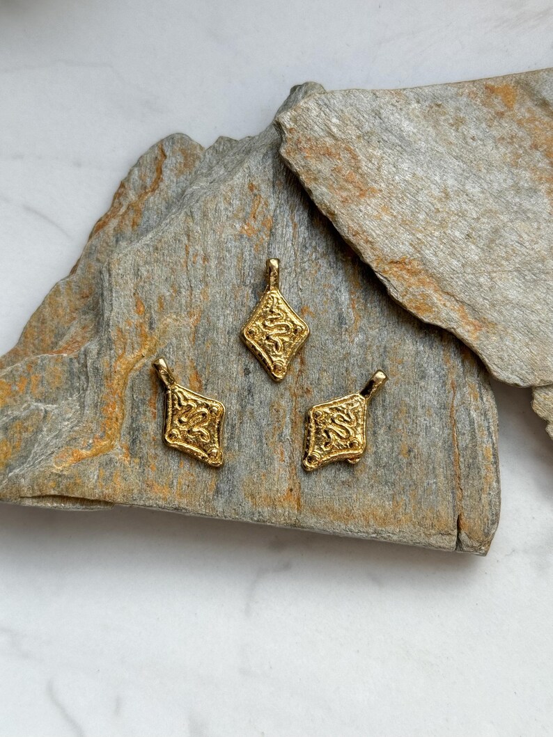Pode incluir: Tr&ecirc;s pingentes dourados e ornamentados, com formato de losango, s&atilde;o exibidos em uma superf&iacute;cie de pedra cinza. Cada pingente apresenta um design intrincado e texturizado, com uma pequena argola no topo para pendurar. Ideal para joias.
