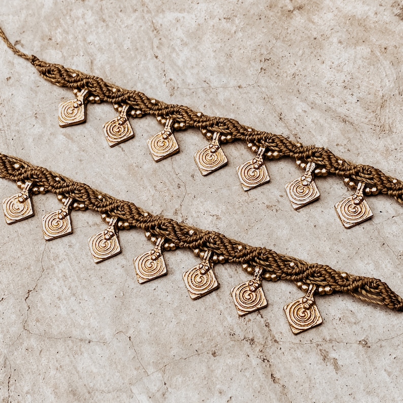 May include: Two brown braided cords with gold metal square charms with a swirl design. The cords are intertwined and the charms hang down.