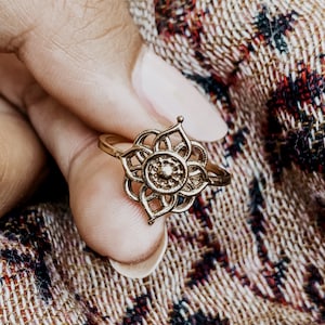 K&ouml;nnte beinhalten: Ein goldfarbener Ring mit einem filigranen, komplizierten Design, das an eine Blume erinnert. Der Ring wird an einem Finger getragen und zeigt die filigranen Details des Designs.