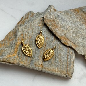 May include: Three gold leaf-shaped pendants with intricate geometric designs. The pendants are arranged on a piece of rough, gray rock.