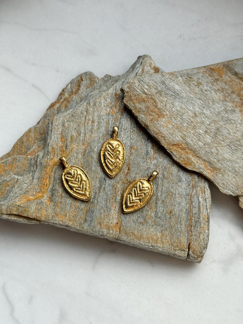 May include: Three gold leaf-shaped pendants with intricate geometric designs. The pendants are arranged on a piece of rough, gray rock.