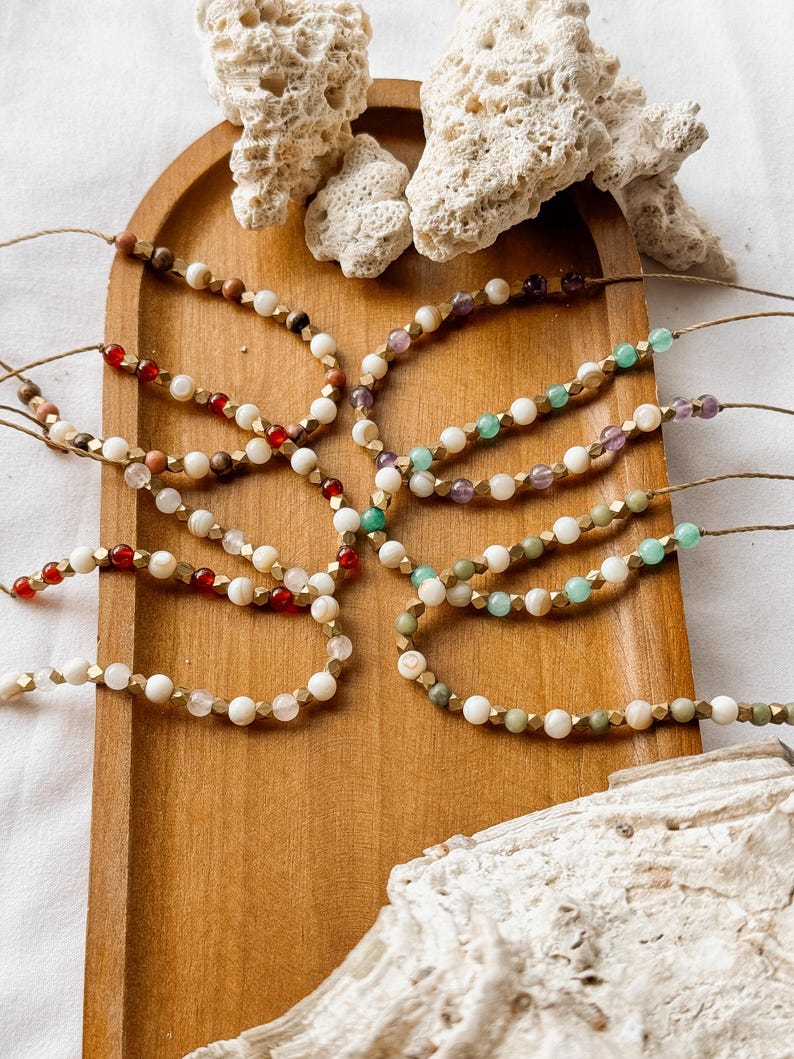 Puede incluir: Una colecci&oacute;n de pulseras de cuentas dispuestas sobre una bandeja de madera, con diversas piedras naturales en tonos rojo, verde, morado y blanco, intercaladas con peque&ntilde;os espaciadores met&aacute;licos dorados y ensartadas en un cord&oacute;n marr&oacute;n.