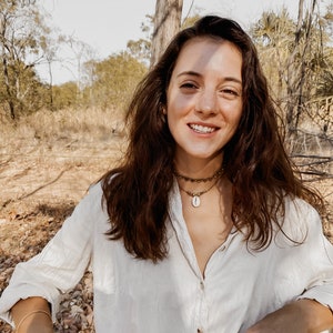 May include: A woman with long brown hair wearing a white button-down shirt and a gold necklace with a shell pendant. She is standing in a wooded area with trees and brown leaves on the ground.