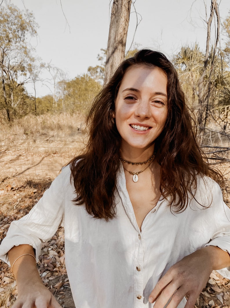 May include: A woman with long brown hair wearing a white button-down shirt and a gold necklace with a shell pendant. She is standing in a wooded area with trees and brown leaves on the ground.
