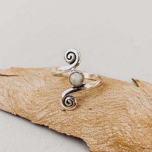 Puede incluir: Un anillo plateado con un diseño en espiral y una pequeña piedra redonda de color claro. El diseño del anillo presenta dos espirales a cada lado de la piedra. El anillo se muestra sobre un trozo de madera flotante.